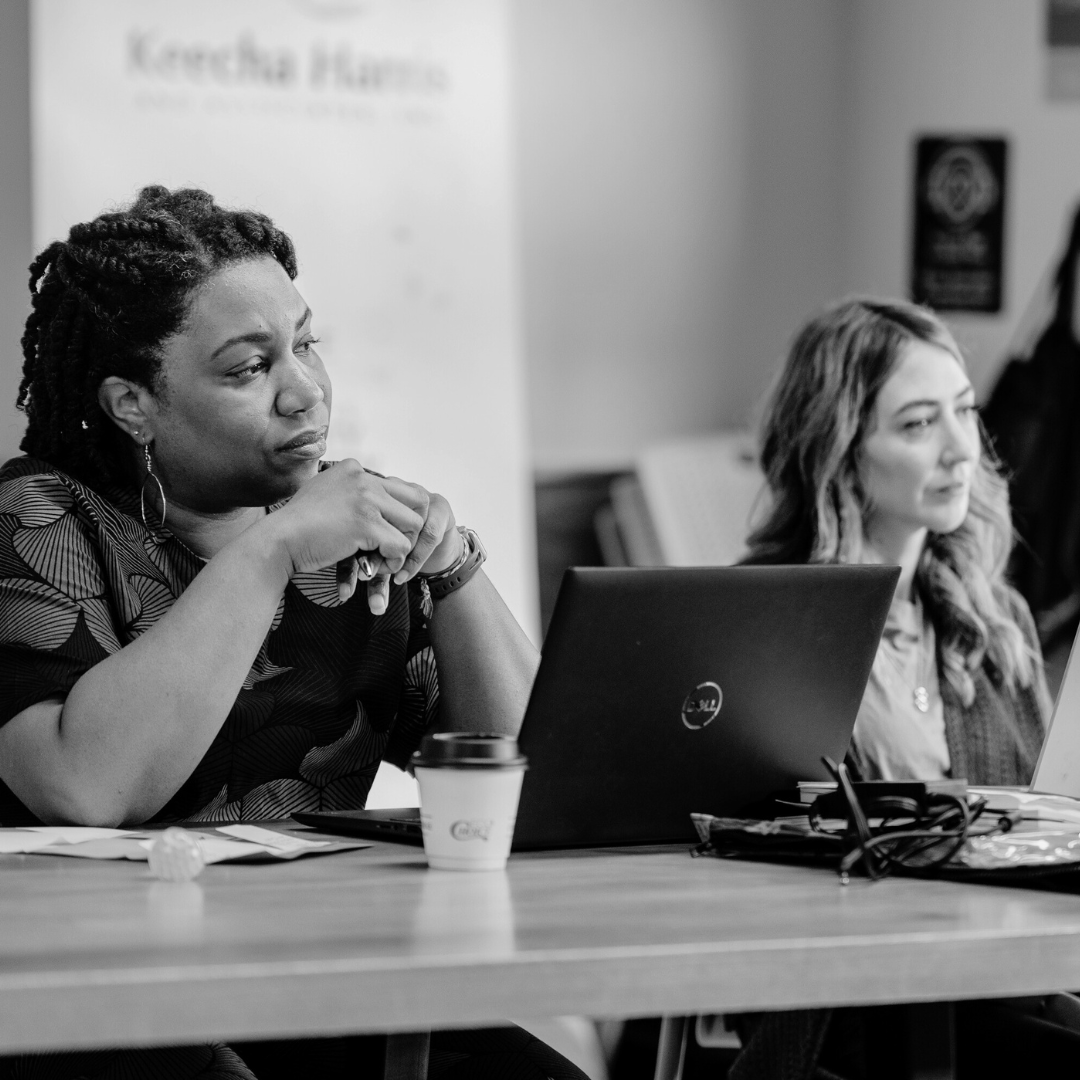 Two team members of Keecha Harris & Associates sitting at a table with laptops listening to a presentation